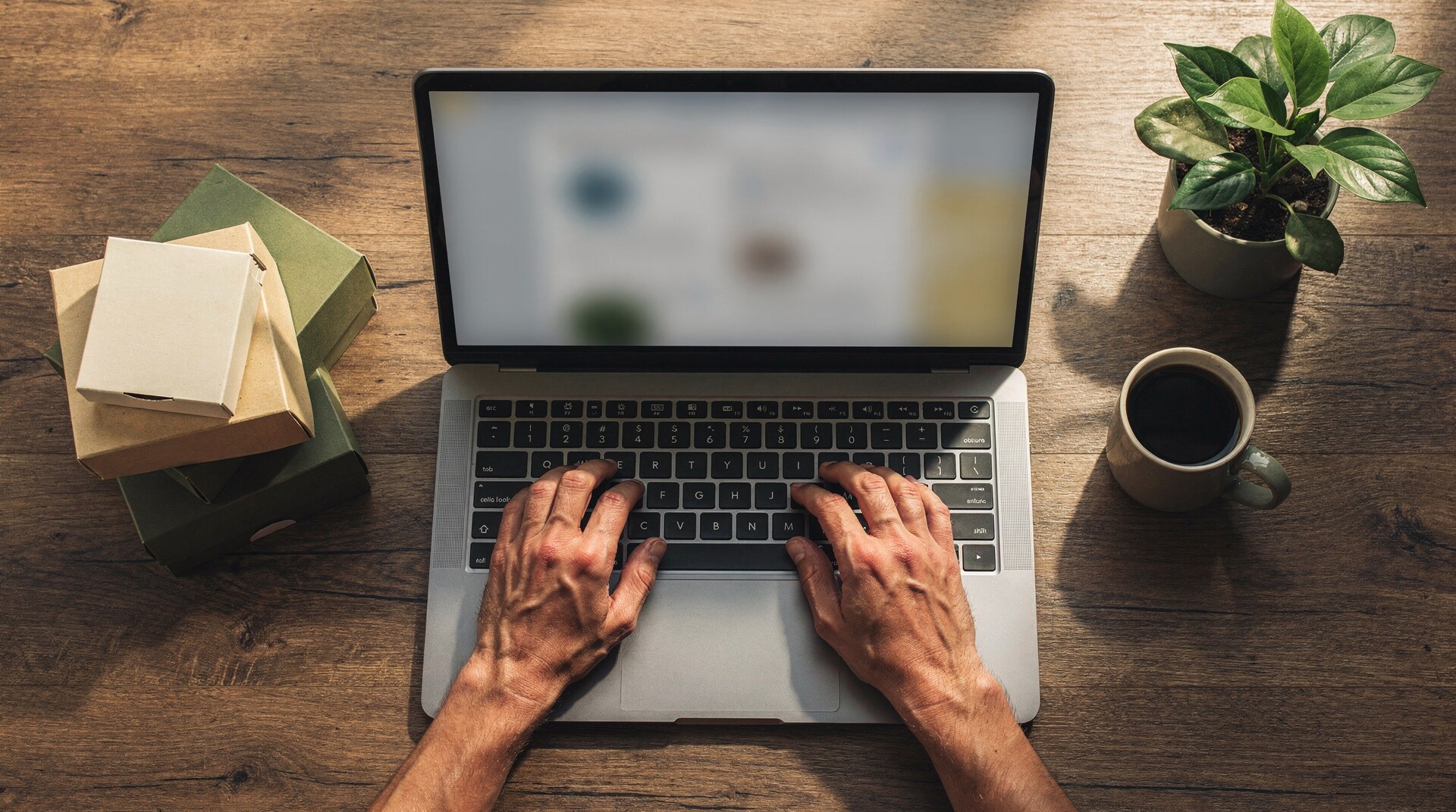 Overhead view of hands typing on a laptop beside stacked subscription box packaging, a plant, and a cup of coffee on a wood desk