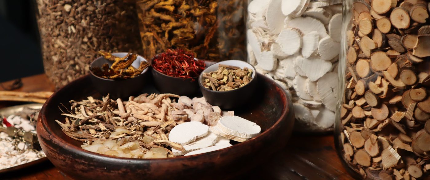 Dried medicinal herbs and roots in wooden bowls beside apothecary jars on a wooden table